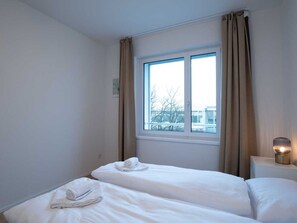 Quarto de hotel com duas camas, uma janela com cortinas, uma mesa de cabeceira com abajur e vista para edifícios.