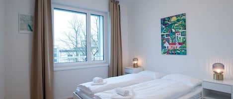 Habitación de hotel con cama, dos mesitas de noche, ventana con cortinas, un cuadro en la pared y una lámpara.