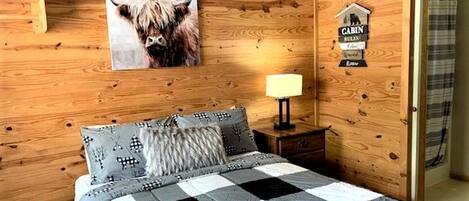 A wooden cabin-style room with a bed, a nightstand, a lamp, and a mounted picture of a cow.