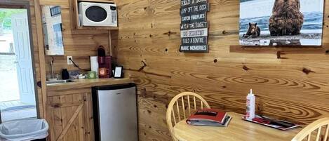 Een compacte keuken met een magnetron, koelkast en houten kastjes. Er is een eethoek met een tafel en stoelen, en een wandklok.