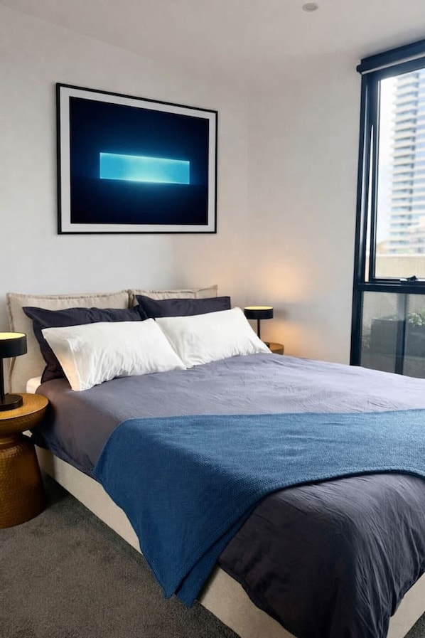 A modern bedroom with a bed, bedside tables, a wall-mounted artwork, and a large window. - Rooftop Infinity Pool & Bay Views (St Kilda)