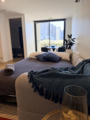 A modern bedroom with a large bed, a view of the city through a window, and a small table with a glass. - Rooftop Infinity Pool & Bay Views Pet Friendly (St Kilda)
