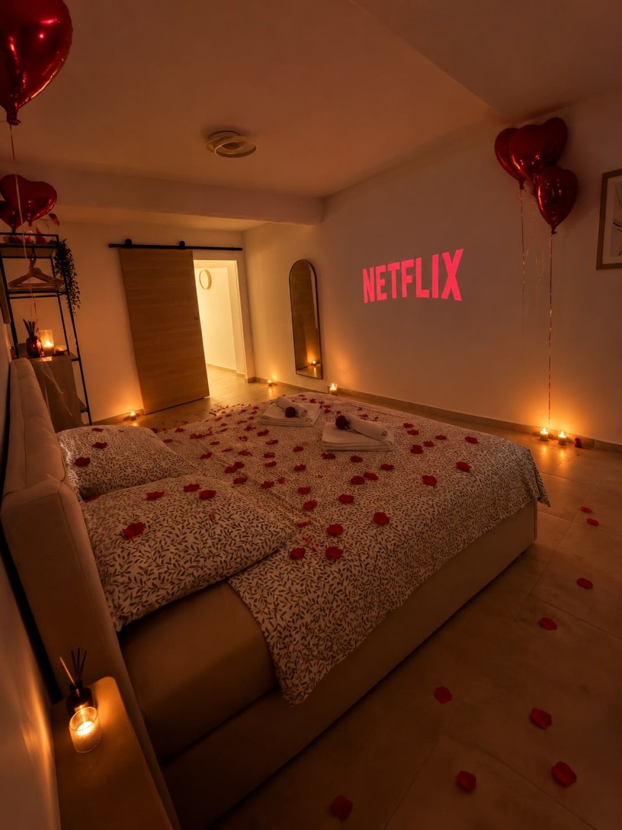 Une chambre à coucher avec un lit, des oreillers, une table de chevet, un téléviseur fixé au mur et des ballons en forme de cœur.