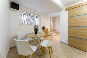 Un comedor con una mesa redonda, cuatro sillas blancas, una planta en maceta y vistas a una escalera que conduce a una puerta de madera.