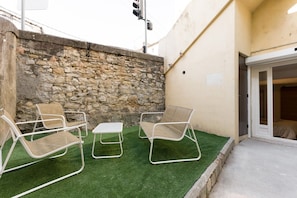 Un pequeño espacio al aire libre con dos sillas y una mesa, un muro de piedra y la entrada de un edificio.