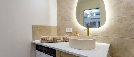Une salle de bain équipée d’un lave-linge, d’un évier avec un robinet doré et d’un miroir à cadre ovale.