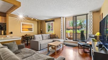 Una sala moderna con sofá, mesa de centro y televisor. Hay un comedor con mesa y sillas, y un balcón con plantas.