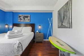 Un dormitorio con una cama, una mesita de noche, una lámpara, una tabla de surf, un cuadro de un mapa mundial y un cuadro de un bosque.