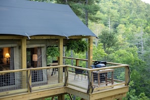 Une terrasse en bois dotée d’un espace salon couvert, d’un barbecue et offrant une vue sur une colline boisée.