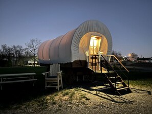 A covered wagon with a staircase leading to an entrance, set in a grassy area with a picnic table and a wooden chair.