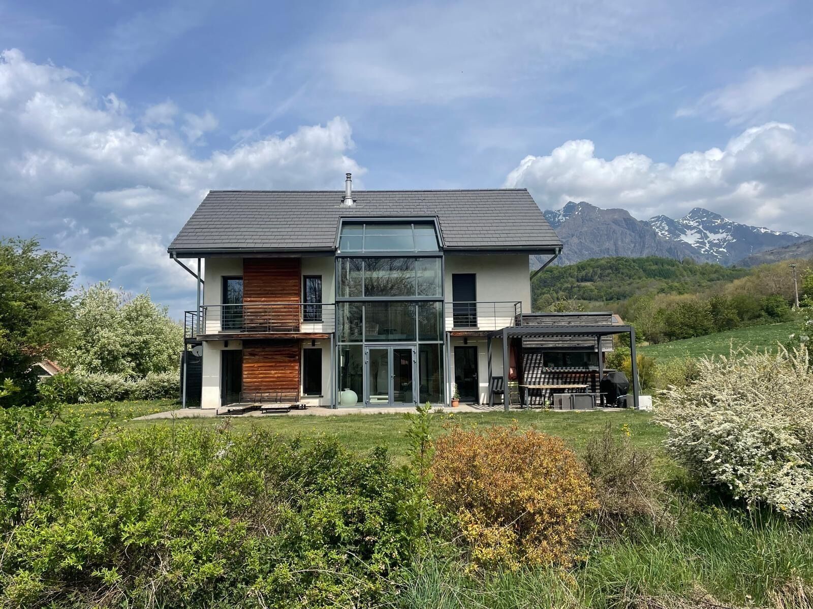 Una casa moderna de dos pisos con un amplio ventanal, una terraza de madera y montañas al fondo.