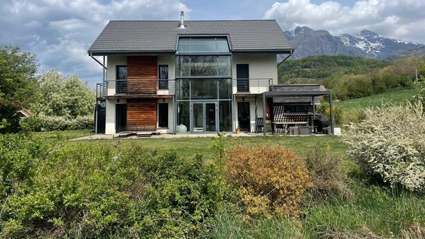 Et moderne hus med to etager, stor glasfacade, træterrasse og et bjerg i baggrunden.