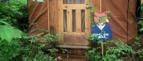 A small, wooden yurt with a door and a sign, surrounded by dense forest.