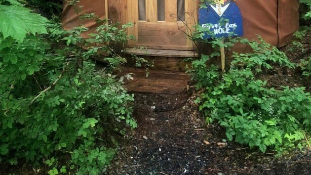 A small, wooden yurt with a door and a sign, surrounded by dense forest.