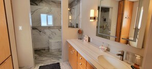 Un baño con ducha de mármol, un espejo grande y tocador con lavabo y toallero.