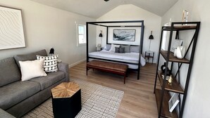 A bedroom with a four-poster bed, a grey sofa, a wooden coffee table, a black ottoman, a patterned rug, and a black metal shelving unit.
