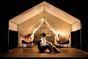 Une tente à l’intérieur de laquelle se trouve une personne assise sur le sol, deux chaises et des guirlandes lumineuses.