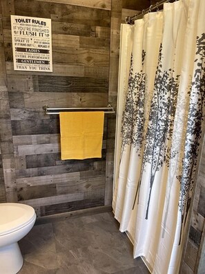 A bathroom with a wooden wall, a toilet, a towel rack with a yellow towel, and a shower curtain with a floral pattern. - Stunning Secluded Dome with Gorgeous Forest Sights in Hermosa, South Dakota (Hermosa)
