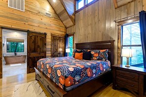 Ein Schlafzimmer in einer Blockhütte mit einem großen Bett, einer Holzwanne und einem Fenster mit Blick auf Bäume.