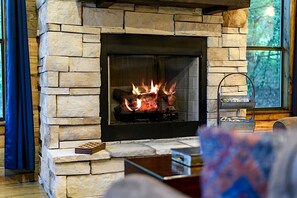 Ein Steinkamin mit Feuer, ein Fenster mit Blick auf Bäume und ein blauer Vorhang.