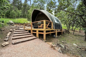 Eine kuppelförmige Hütte mit einer Holzdachterrasse und einem Stuhl, umgeben von einem Wald.