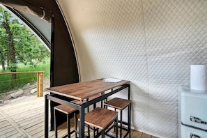A tent with a wooden table and two stools, a white quilted wall, and a view of greenery outside.