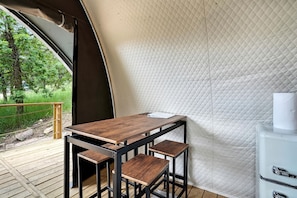 Een tent met een houten tafel en twee krukken, een witte gewatteerde wand en uitzicht op groen buiten.