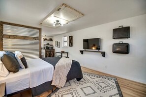 Une chambre à coucher avec un lit, une table de chevet, une télévision, des étagères murales et, au fond, un coin cuisine.