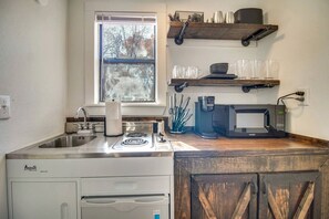 A compact kitchen with a stove, sink, and microwave. There is a window above the sink, and shelves above the counter holding dishes and glasses. - Cozy Studio Retreat with Scenic 5-Acre Views, Near City Lake & Cherokee Casino in Gentry, Arkansas (Gentry)