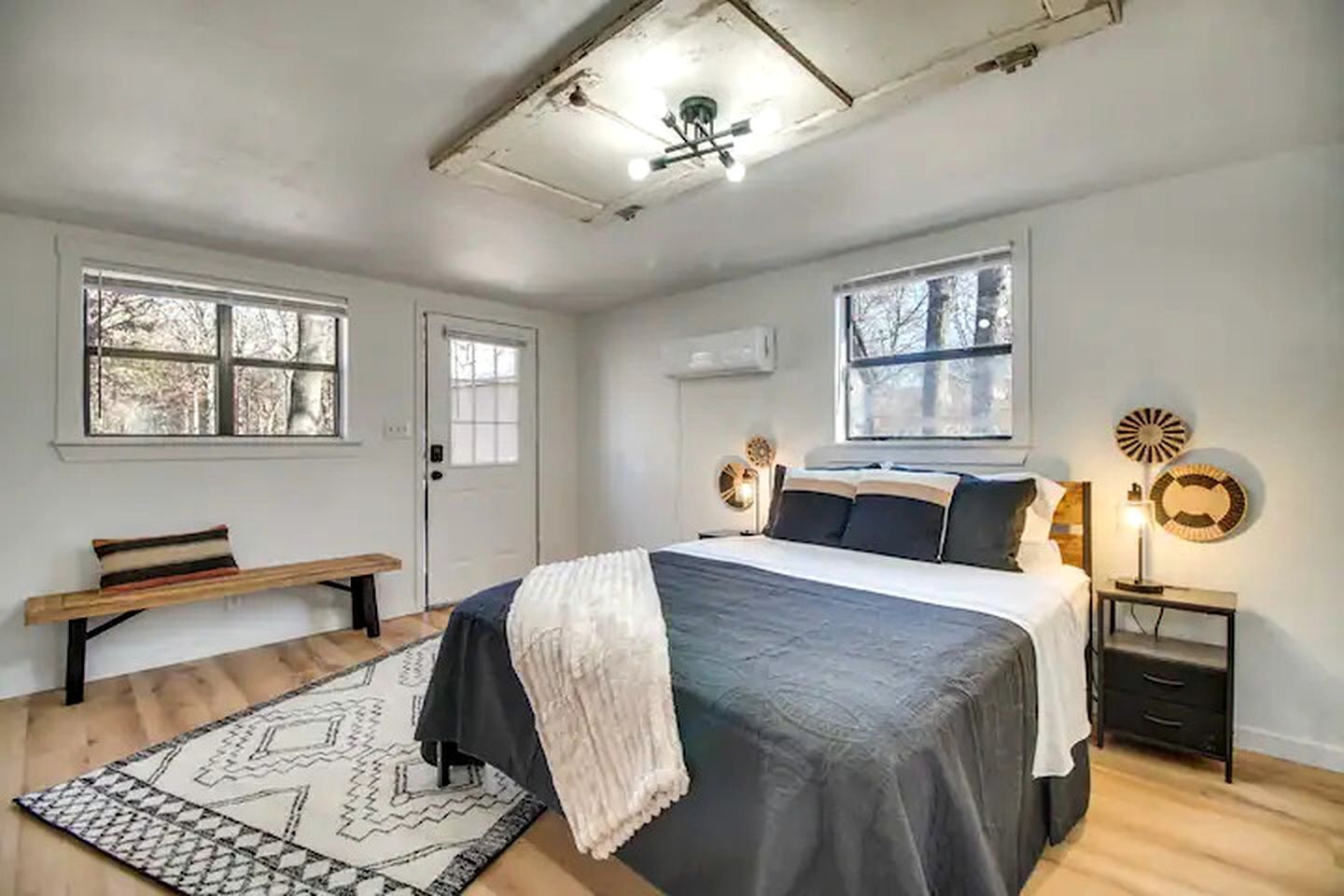 Un dormitorio con una cama, mesitas de noche, un banco, una alfombra estampada y dos ventanas.