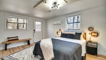 Un dormitorio con una cama, mesitas de noche, un banco, una alfombra estampada y dos ventanas.