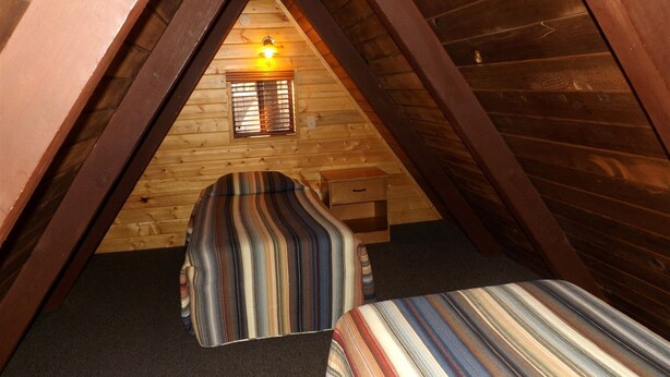 Un dormitorio abrigado en el ático, con techo inclinado, paredes de madera, un ventanal con persianas y dos camas a rayas.
