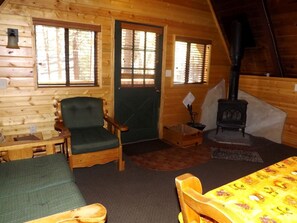 Ein gemütliches Blockhaus-Interieur mit Holzausbau, einem grün gepolsterten Sessel, einem Tisch mit gelber Tischdecke, einem Kamin und einem Fenster mit Jalousien.