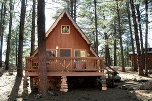 Una cabaña con terraza de madera, base de piedra y techo triangular, rodeada de árboles.