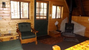 Interior acogedor de una cabaña con detalles de madera, un sillón tapizado en verde, una mesa con mantel amarillo, una chimenea y una ventana con persianas.