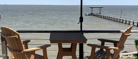 A wooden deck with two Adirondack chairs and a table under a green umbrella, overlooking a body of water and a pier.
