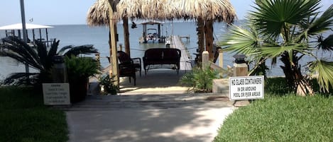 A pathway leading to a thatched structure with outdoor seating, a dock, and a sign prohibiting glass containers in the pool area.
