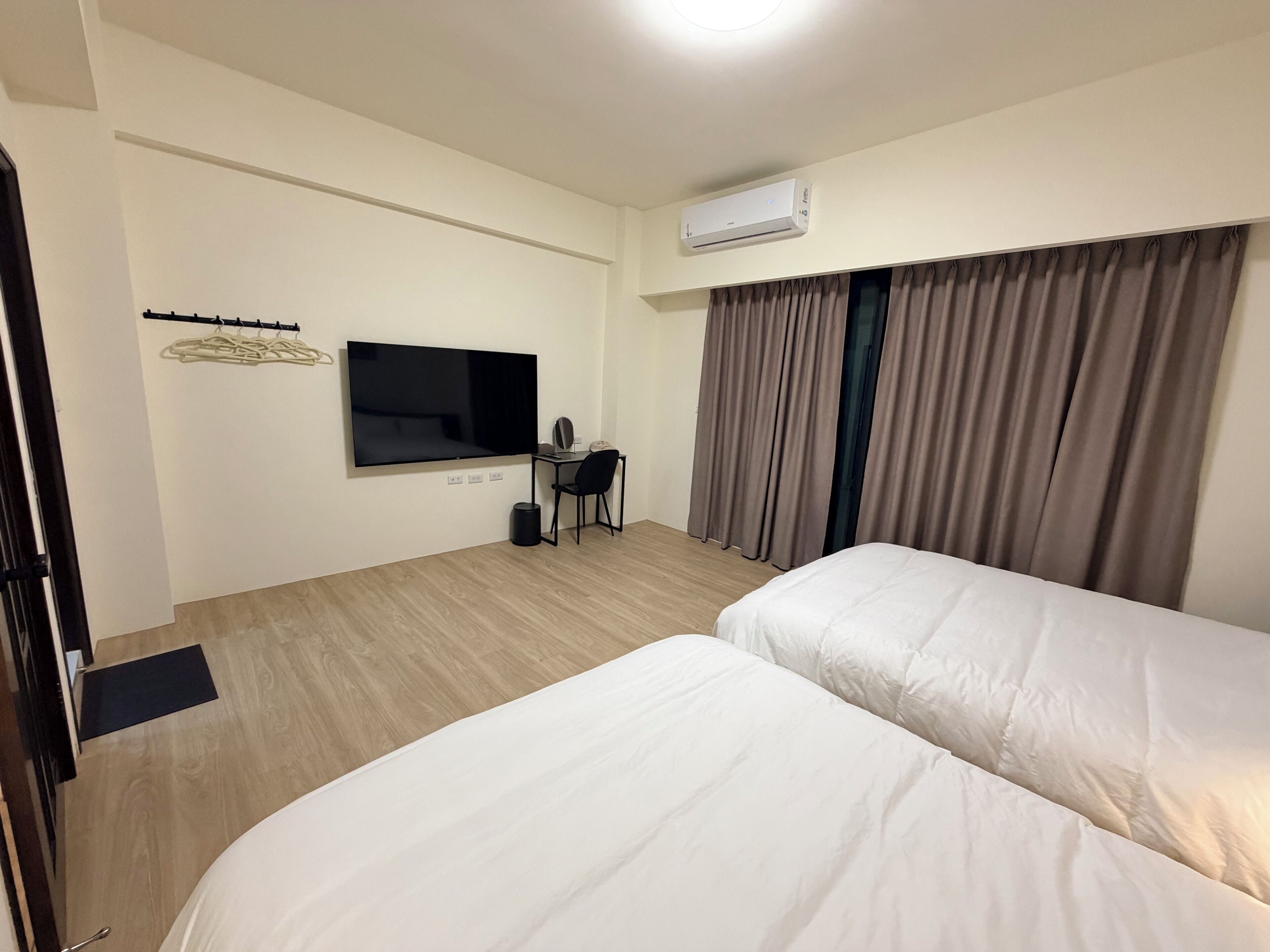 Une chambre d’hôtel avec deux lits, une télévision, un bureau avec une chaise, un porte-manteau et une fenêtre avec des rideaux.
