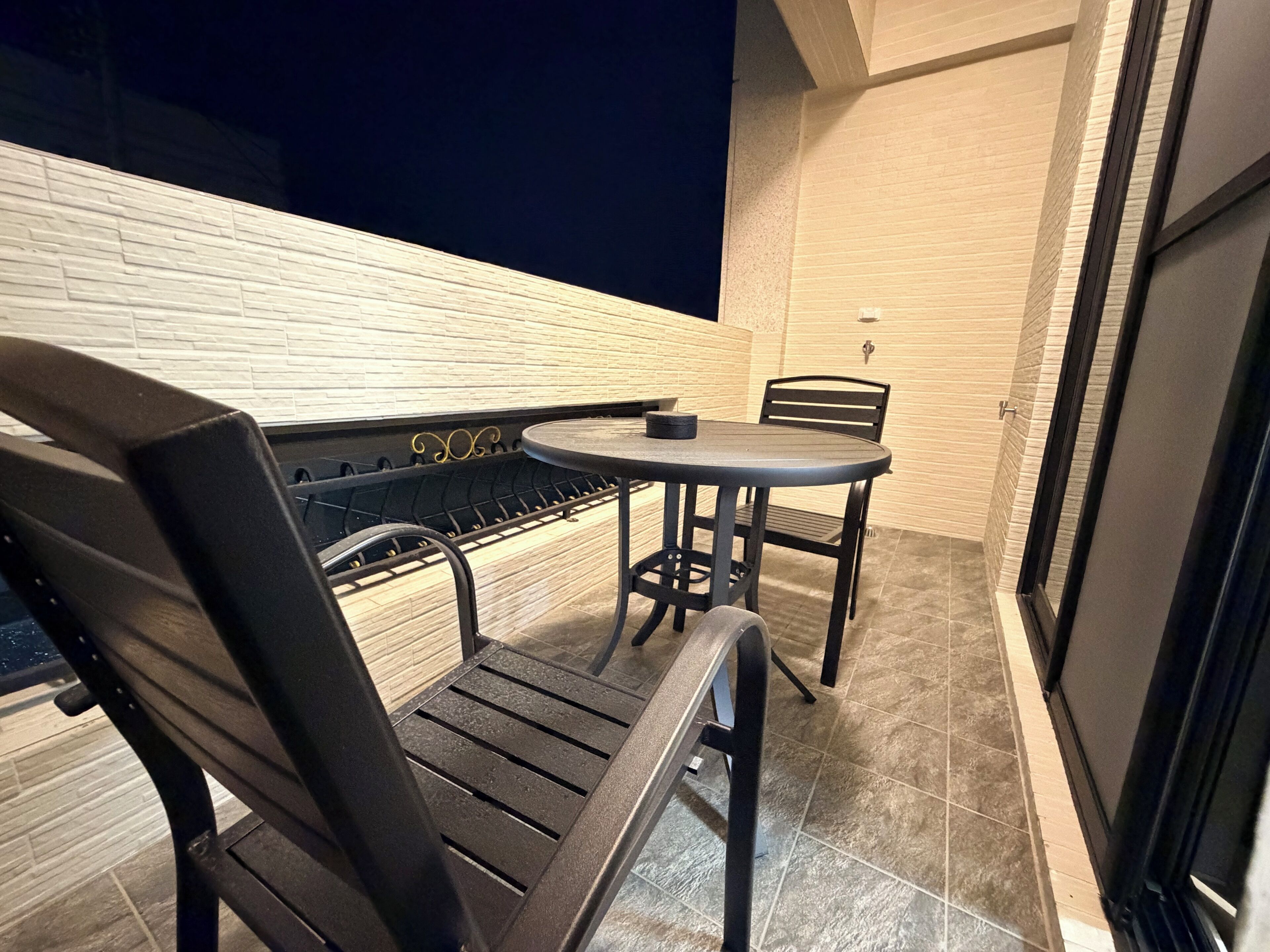 A small outdoor seating area with a table and two chairs, a tiled floor, and a white brick wall.