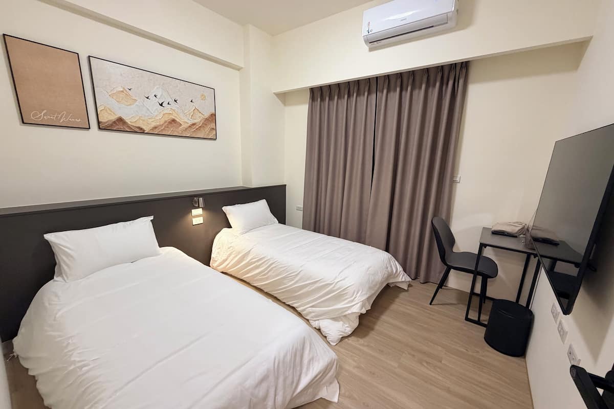 Ein Hotelzimmer mit zwei Betten, einem Schreibtisch mit Stuhl, einer Klimaanlage und zwei Wandbildern.