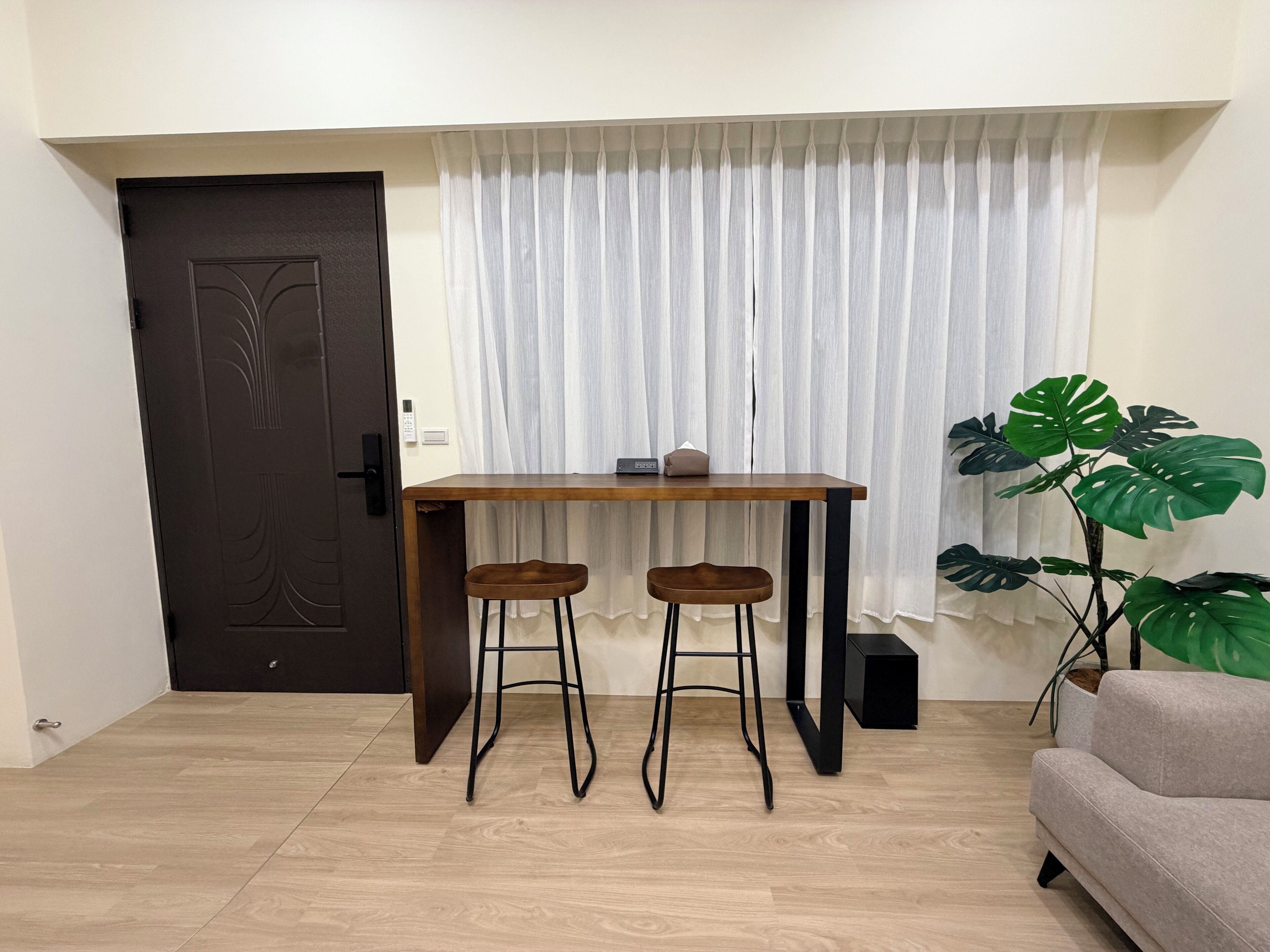A room with a wooden table, two stools, a dark door, a white curtain, a potted plant, and a sofa.