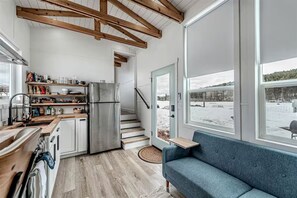 A modern kitchen with a stainless steel refrigerator, wooden beams, a built-in oven, and a window overlooking a snowy landscape. - Gorgeous and Secluded Tiny House in Big Sky Country with Access to Monture Creek, Montana (Ovando)