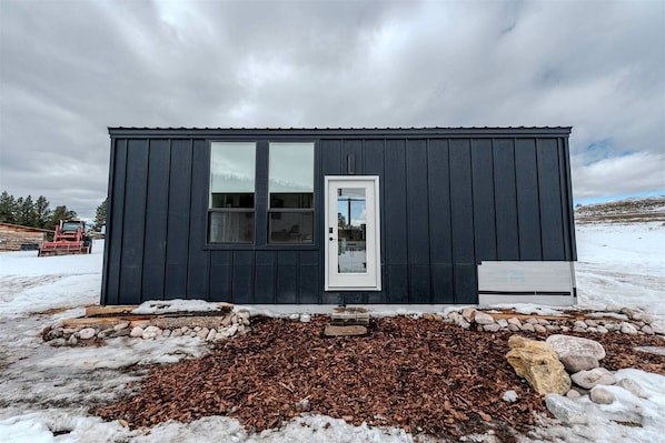 A small, dark-colored cabin with a single door and two windows, surrounded by a snowy landscape.