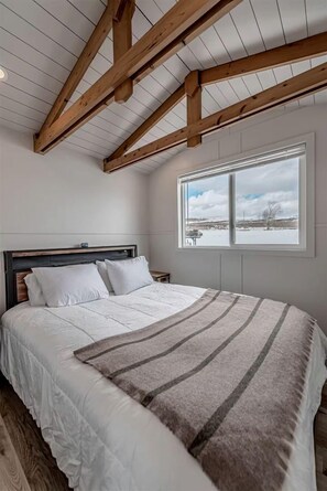 A bedroom with a wooden ceiling, a large bed with white bedding, a window with a view of a snowy landscape, and a bedside table. - Gorgeous and Secluded Tiny House in Big Sky Country with Access to Monture Creek, Montana (Ovando)