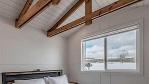 Un dormitorio con techo de madera, una cama grande con ropa blanca, una ventana con vistas a un paisaje nevado y una mesita de noche.