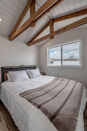 Une chambre avec un plafond en bois, un grand lit avec une literie blanche, une fenêtre donnant sur un paysage enneigé et une table de chevet.