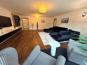A modern living room with a black sectional sofa, a white coffee table, patterned armchairs, a flat-screen TV mounted on the wall, and a large window.