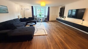 En moderne stue med en hjørnesofa, et sofabord, et fjernsyn og et stort vindue med gardiner.