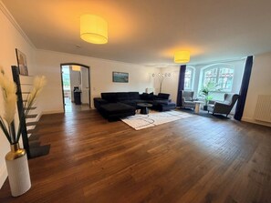 A spacious living room with hardwood flooring, a black sectional sofa, a coffee table, and two pendant lights.