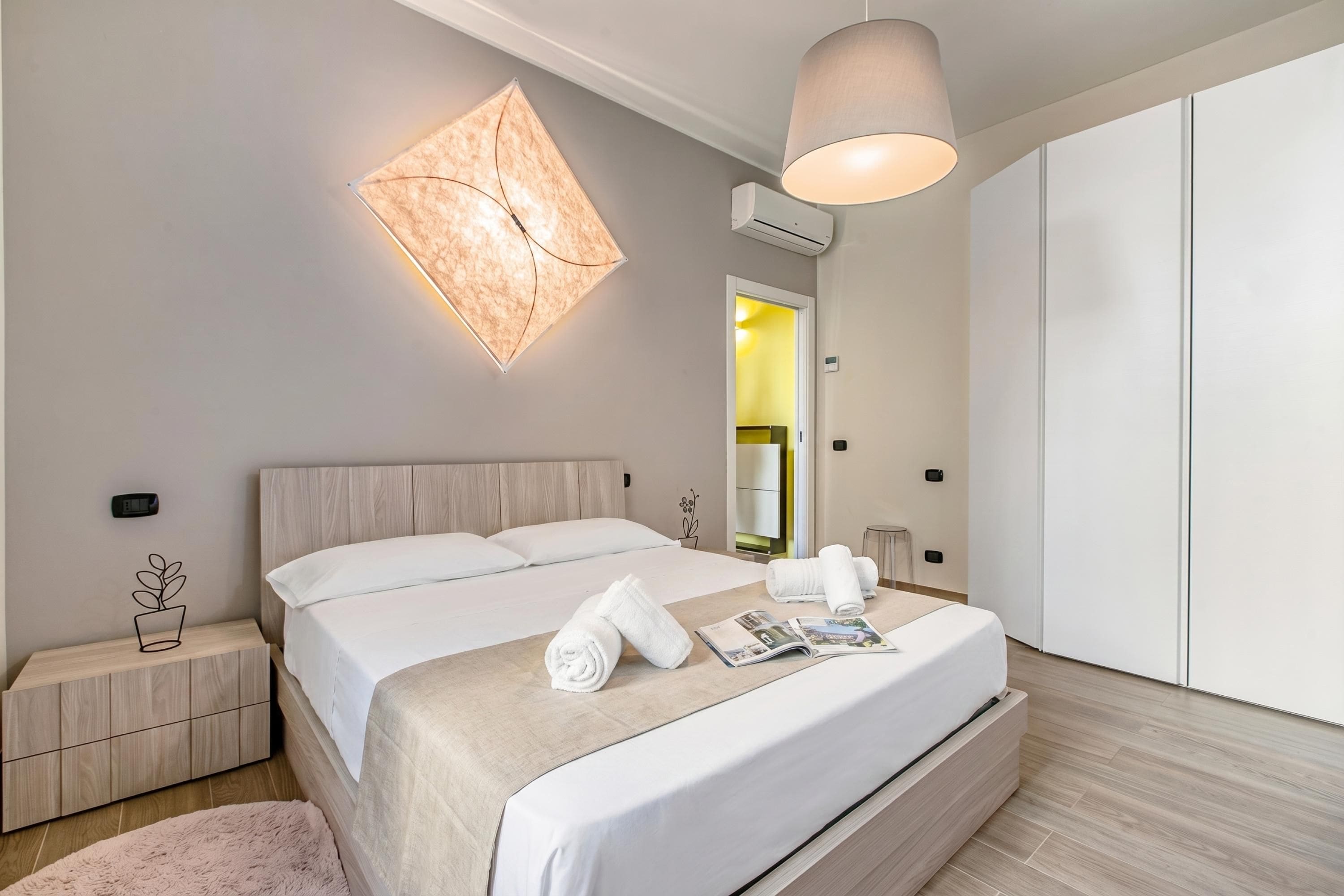 Quarto de hotel moderno com uma cama grande, mesas de cabeceira, luminária fixada na parede e um guarda-roupa.